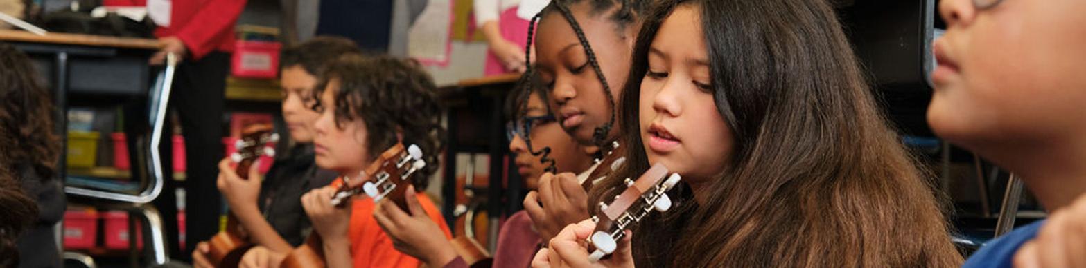 Six elementary-aged children of diverse backgrounds sit on a carpeted classroom floor, each playing a ukulele. Five adults stand behind them, observing the music lesson.